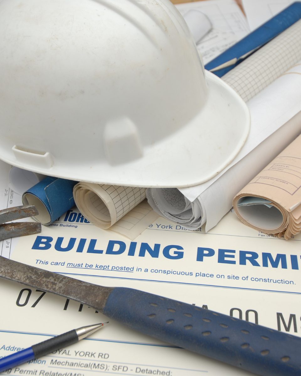 A California building permit card posted on a construction site alongside rolled blueprints, a hard hat, and framing tools