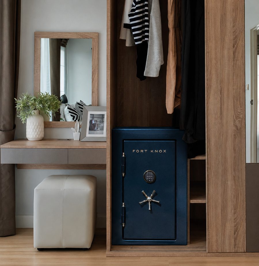 A navy Fort Knox compact safe installed at the base of a fitted bedroom closet beside hanging clothes, a dressing table, and a mirror