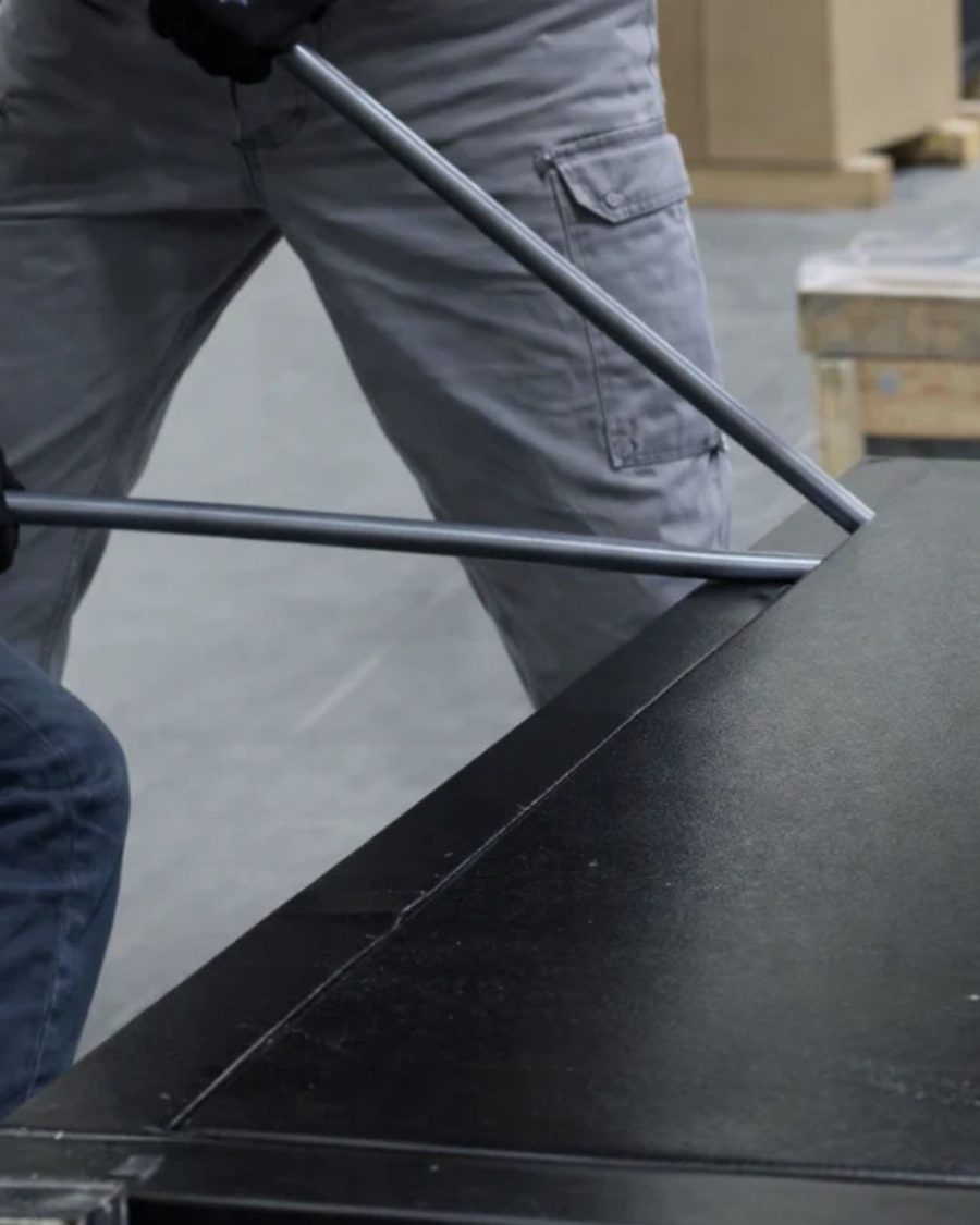 A gloved intruder using a pry bar against the seam of a black steel safe body on a workshop floor, testing its burglary resistance