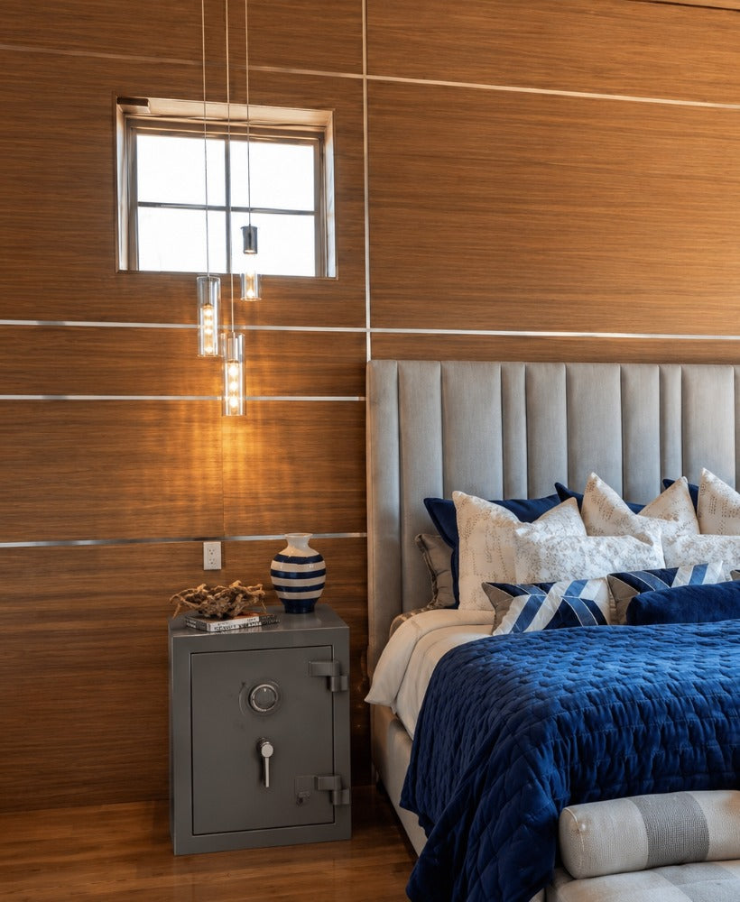 A gray bedside quick-access safe mounted at nightstand height beside a bed, positioned for rapid home defense access in the bedroom