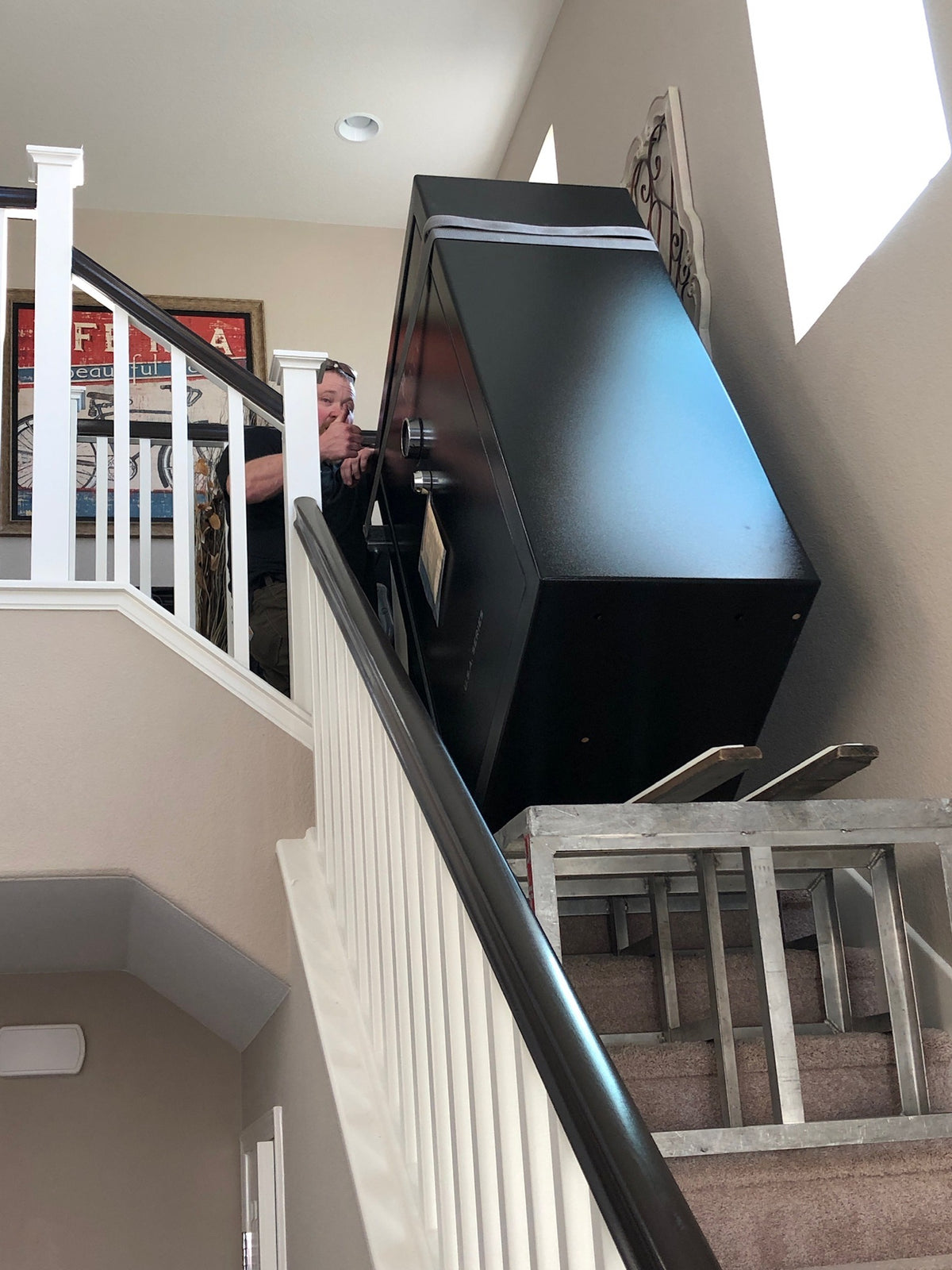 A crew member guiding a large safe strapped to a stair-climbing dolly up an interior residential staircase