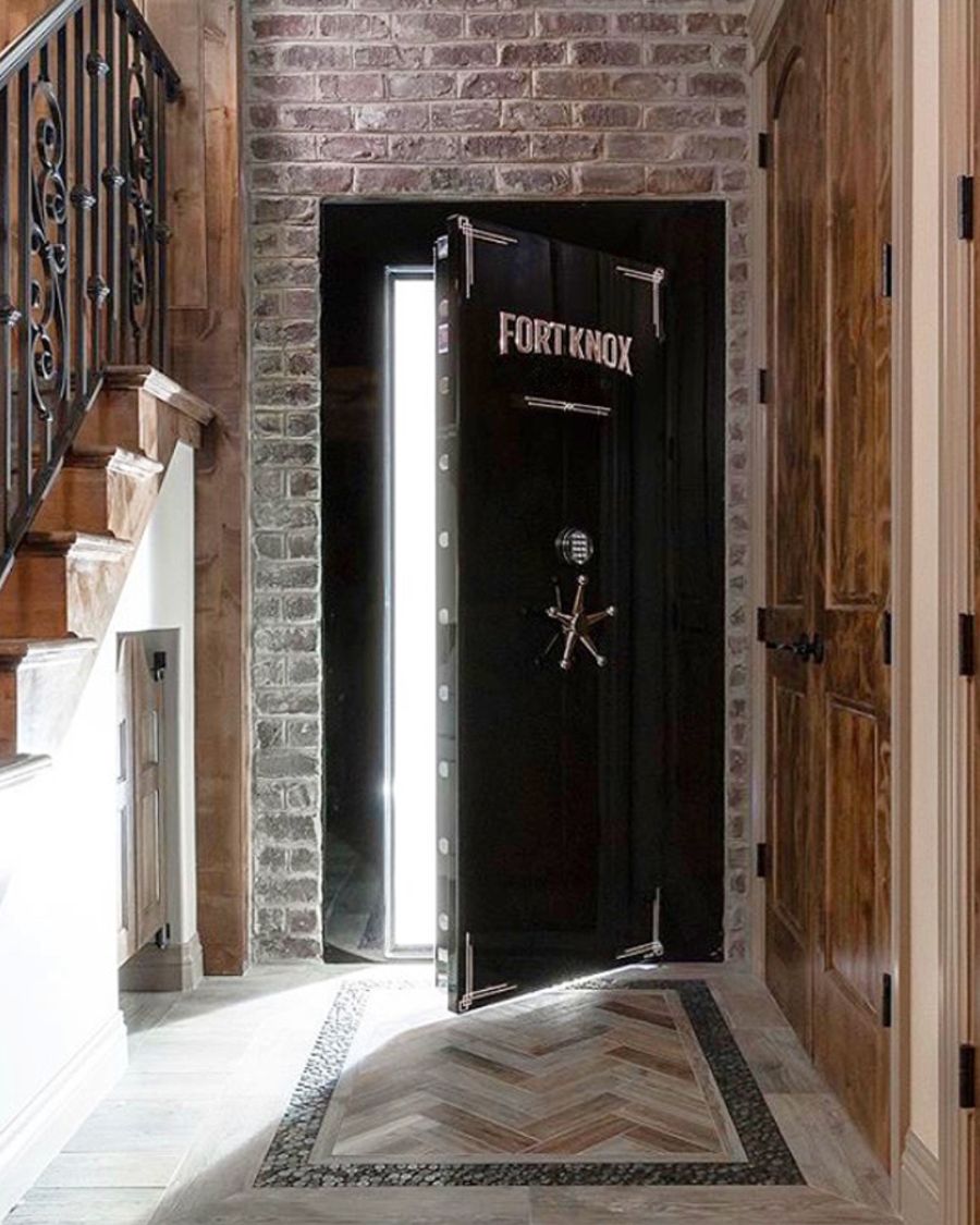 A finished residential vault room entrance: a black Fort Knox vault door standing open in a brick-and-wood alcove beside a staircase, with a herringbone tile inlay in the floor