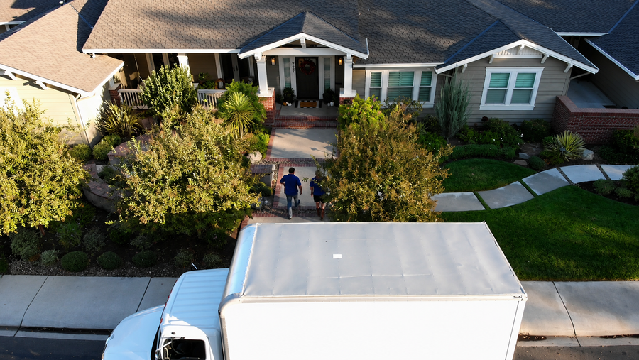 Aerial view of an unmarked white truck parked at the curb of a craftsman home as a crew walks up the front path