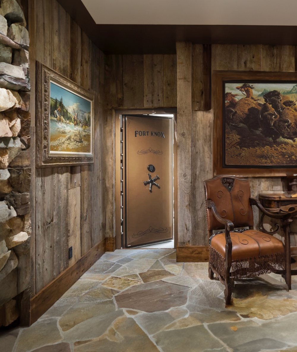 A Fort Knox vault door set into a finished rustic vault room with a rock wall, reclaimed wood paneling, framed western art, and a leather chair