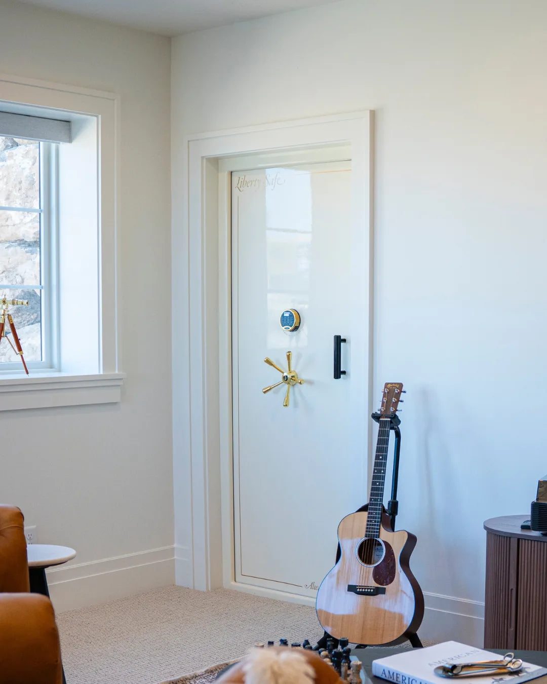 A cream Liberty vault door with a gold five-spoke handle set into a bright, finished residential room with a window and an acoustic guitar